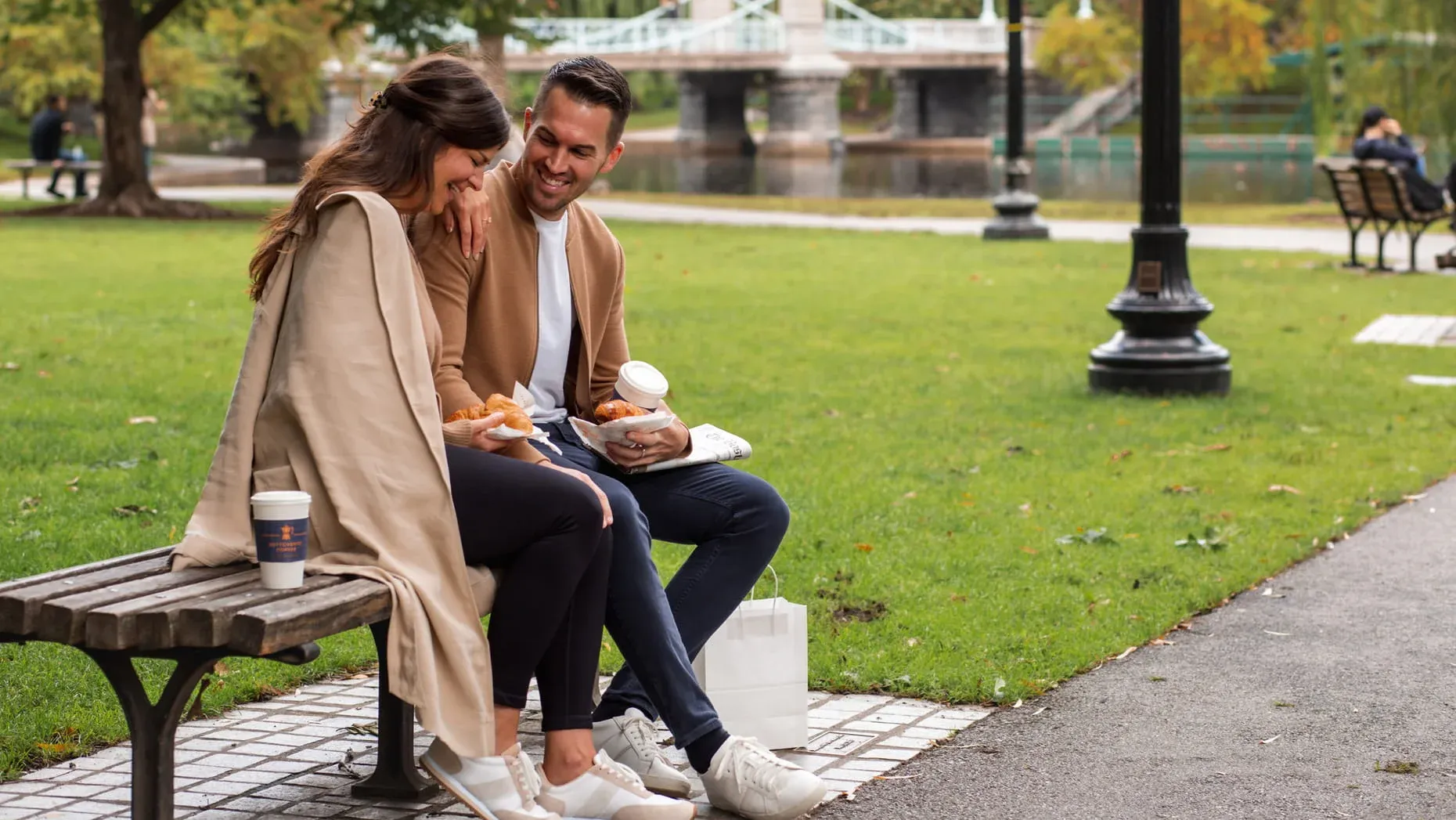 Couple Sitting on Bench In Public Garden With Coffee In Hands